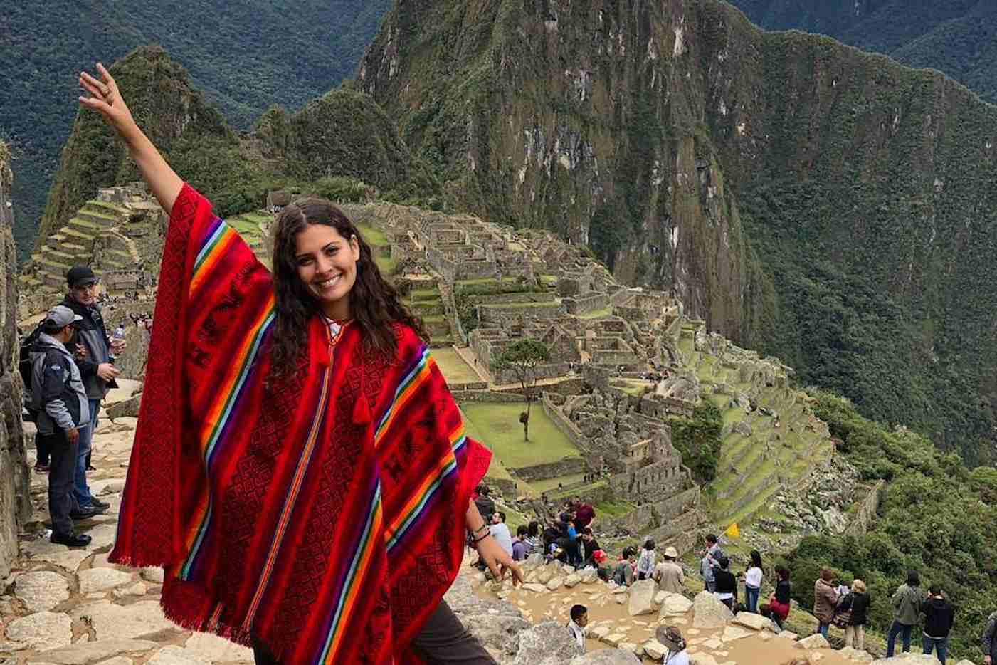 Mujer sonriente con poncho rojo tradicional posando con un brazo alzado frente a las ruinas de Machu Picchu y las montañas andinas.